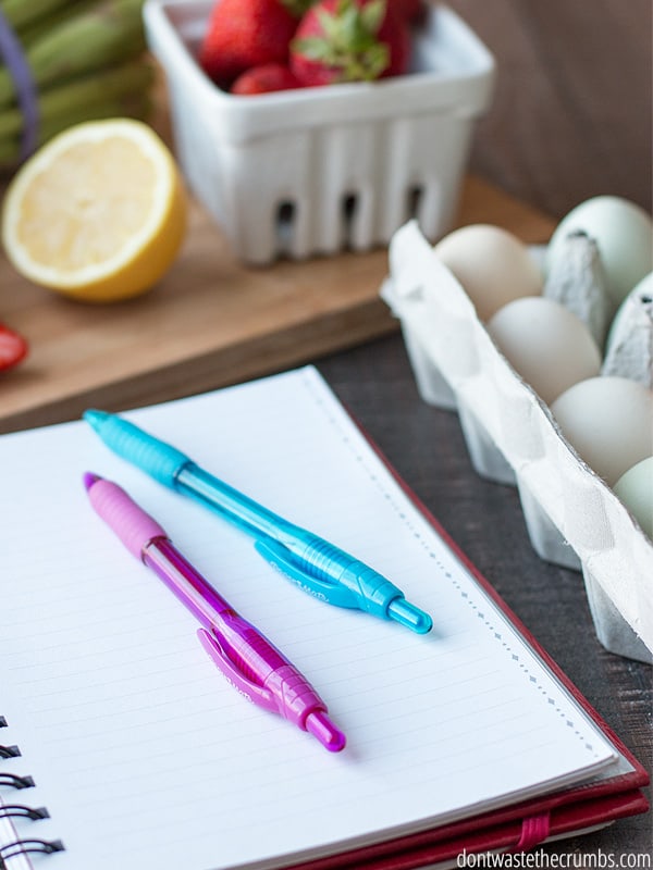 Paper notebook on the table with a pink and blue pen on top, with a carton of eggs to the right of it, and a slice of lemon and a carton of strawberries behind it, ready for planning a freezer cooking day. 