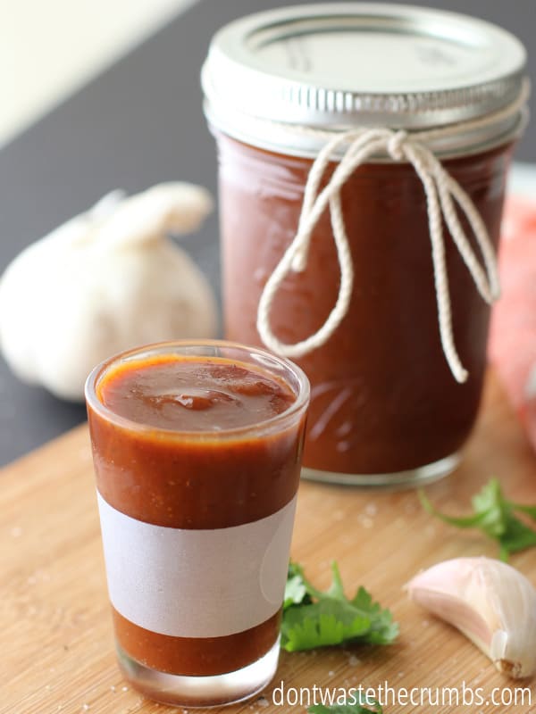 Large mason jar filled with homemade BBQ sauce, kitchen towel with wood spoon on top, garlic clove, cilantro, and a glass cup of BBQ sauce.