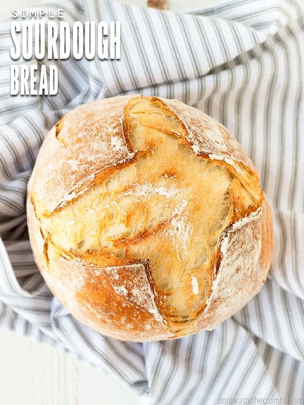 A freshly baked crusty sourdough boule sitting on top of a striped tea towel. The text overlay reads "Simple Sourdough Bread."