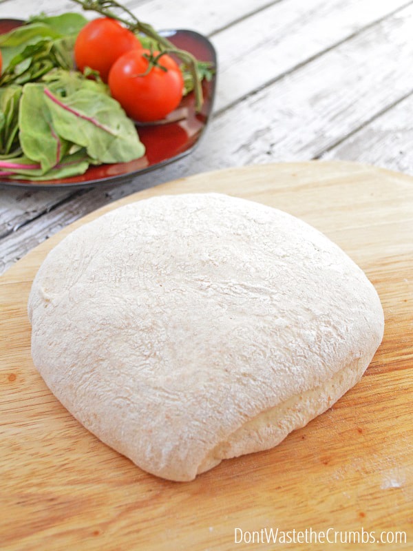 Freshly risen homemade pizza dough sits atop a wooden cutting board, on top of a weathered slatted wooden table. There is a small red plate with a bunch of fresh basil leaves and two vine ripened tomatoes.