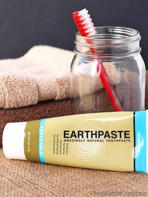 Earthpaste and mason jar with toothbrush in it. Folded towels behind items.