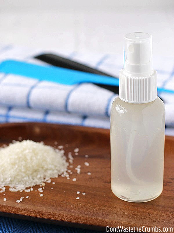 In the foreground, a wooden tray holds a plastic spray bottle of a liquid to naturally condition hair, along with a small pile of sea salt. In the background, two combs lie on a folded towel