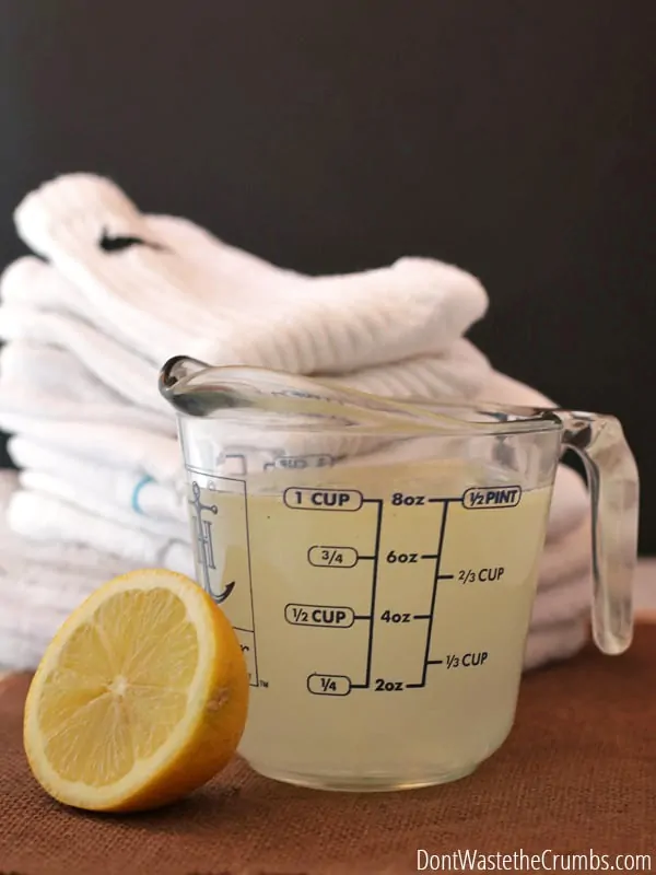 A stack of white clothing articles behind a measuring cup full of bleach alternative and a sliced lemon.