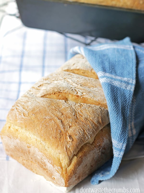 A loaf of bread wrapped in a blue kitchen towel. Making your own bread is a great way to save money!