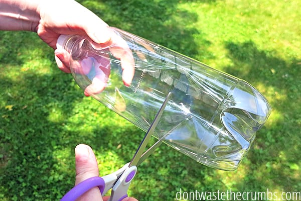 Hands holding and cutting bottom of plastic bottle with scissors.