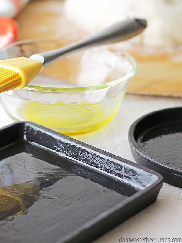 Pictured is a glass bowl with olive oil and a basting brush laying on top of the bowl. There are two cast irons and they have been lightly coated with olive oil.