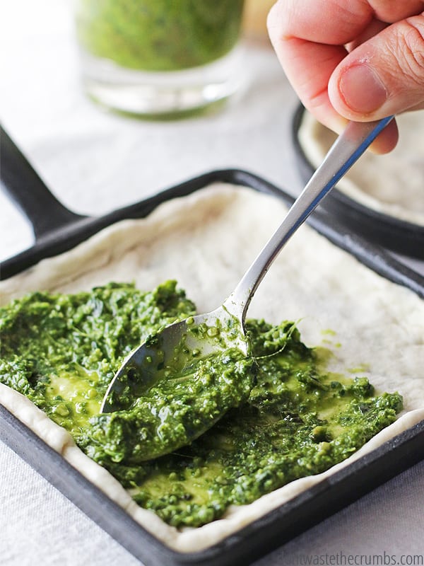 This cast iron skillet pizza dough is laid out onto a cast iron and there is carrot pesto being spread throughout with a spoon.