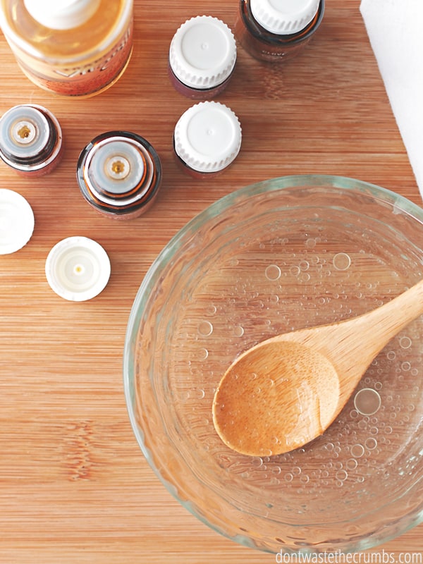 A clear bowl filled with Vitamin E oil, essential oil drops and a wooden spoon inside stirring it. There are five bottles of essentials oils, and vitamin E oil.