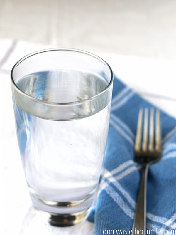 A clear glass of water on top of a blue and white kitchen towel. There is a fork face-up on the towel beside the glass of water. 