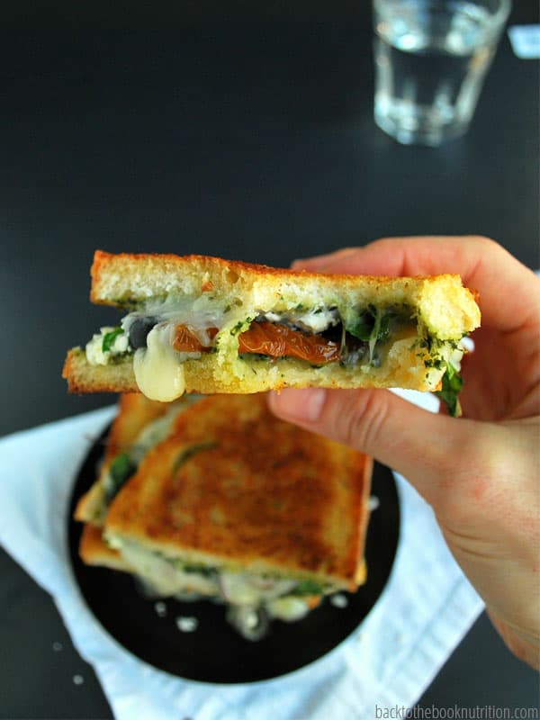 Up close view of a hand holding half of a Mediterranean grilled cheese sandwich and there is a whole sandwich on a black plate in the background.