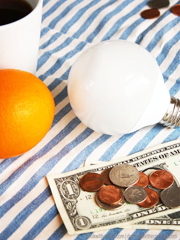 Two dollar bills and a collection of change sit next to a light bulb and an orange.