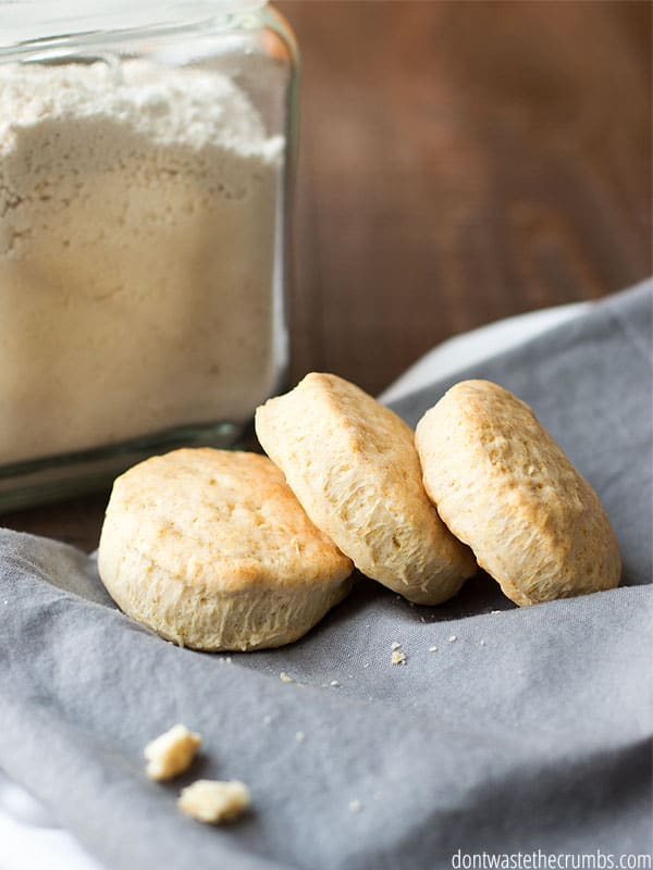 Three homemade biscuits on top of a cloth with a large jar of homemade Bisquick behind it