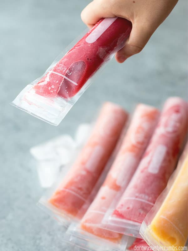 A small hand grabs on to a red ice pop, with orange, red, and yellow ice pops shown below.
