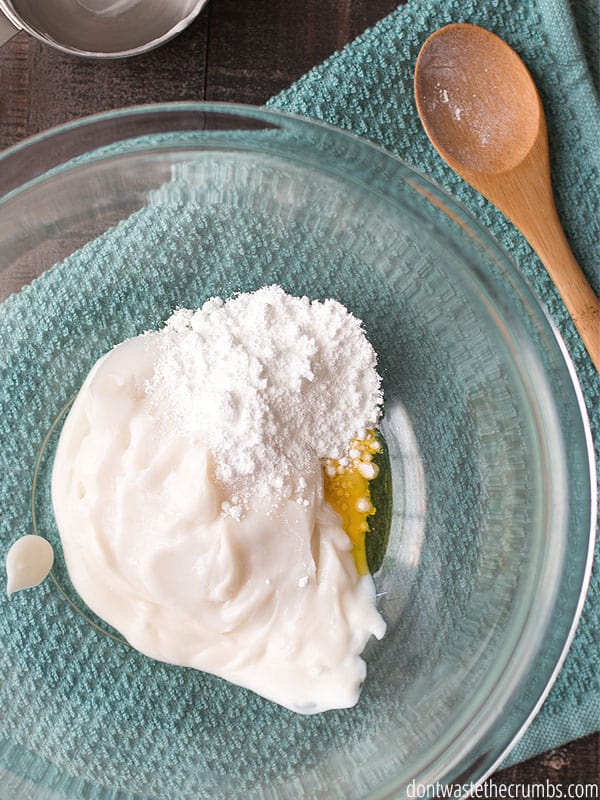 Ingredients for homemade sunscreen in a large glass bowl on top of a dish towel. There is a wooden spoon nearby.