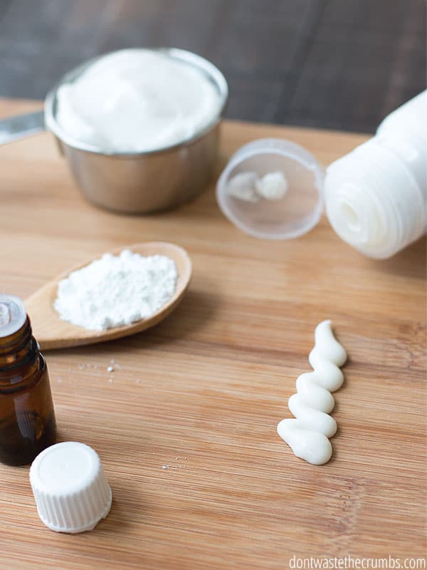 Measuring cup with lotion, wood spoon with powder, squeezable tube, and a small amount of homemade sunscreen are displayed on a wood cutting board.