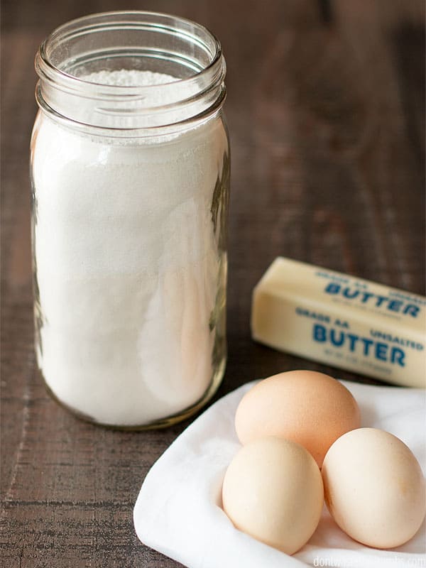 A mason jar is filled with dry cake mix ingredients: flour, sugar, salt, and baking powder. Next to the jar sit 3 eggs and a stick of butter.