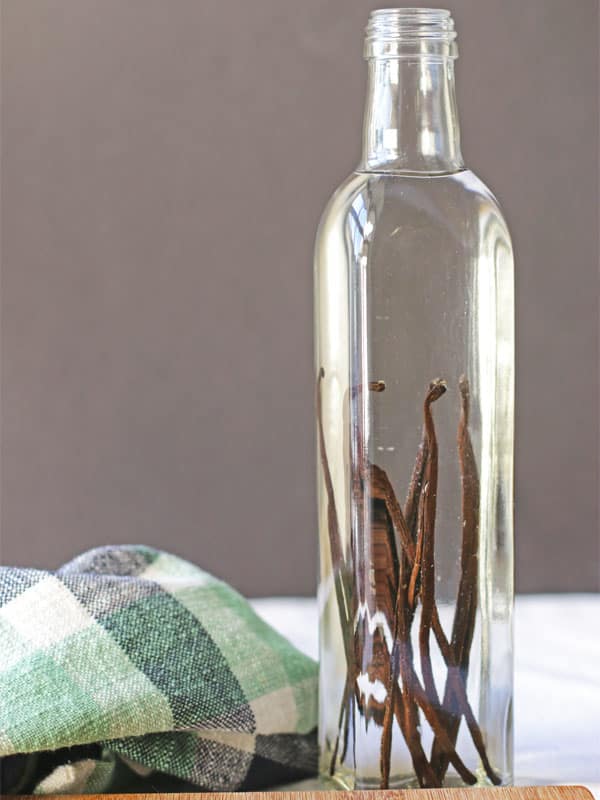 A glass bottle with several vanilla beans is getting ready to be made into homemade vanilla extract. Beside the glass bottle is a green plaid tea towel.