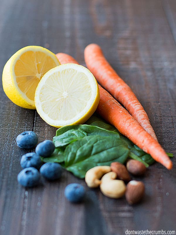 A sliced lemon by two whole carrots, some juicy blueberries, fresh spinach, cashews, and almonds. Leftover scraps from other recipes can make great meals. 