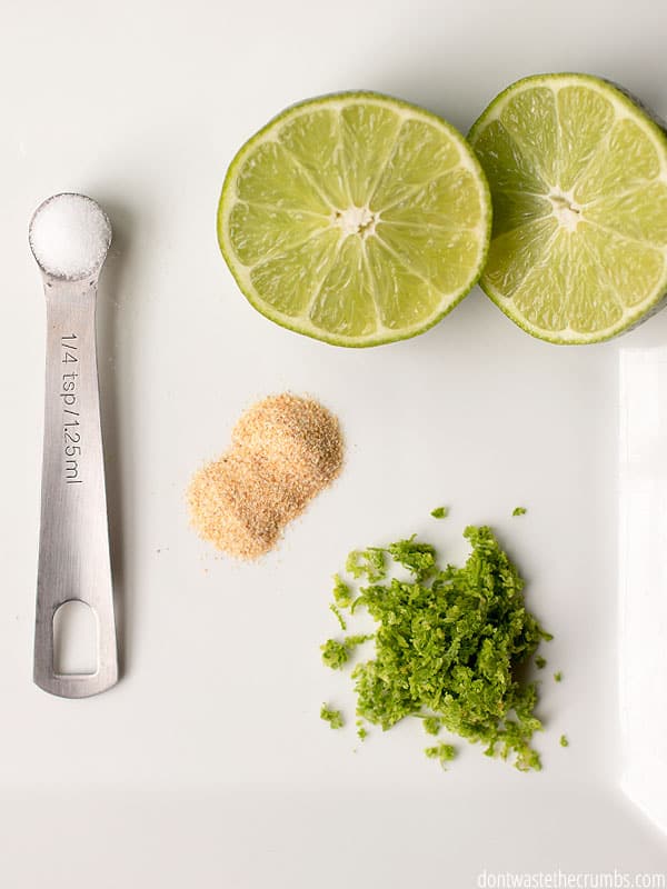 Several ingredients for lime crema, including salt in a measuring spoon, garlic powder, halved limes, and chopped cilantro