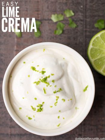 A close-up of a bowl of lime crema on a wooden surface, with a lime slice and cilantro sprinkled nearby