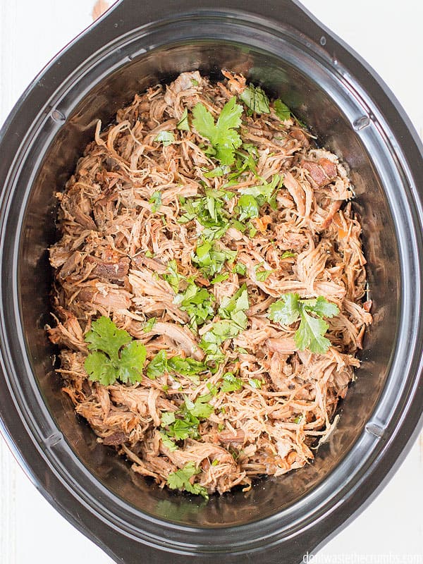 A black oval crockpot with slow cooker carnitas freshly cooked. Cilantro is sliced and sprinkled on top. 