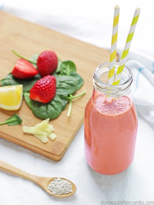 Bentonite clay smoothie in a glass jar. There is strawberries, lemon, spinach, and fresh ginger are on a cutting board and a wooden spoon with bentonite clay. Those are all ingredients for bentonite detox smoothie!
