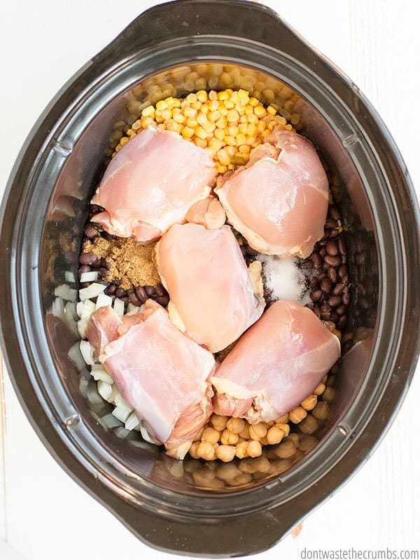 Raw chicken. garbanzo beans, spices, and black beans are in a slow cooker pot.