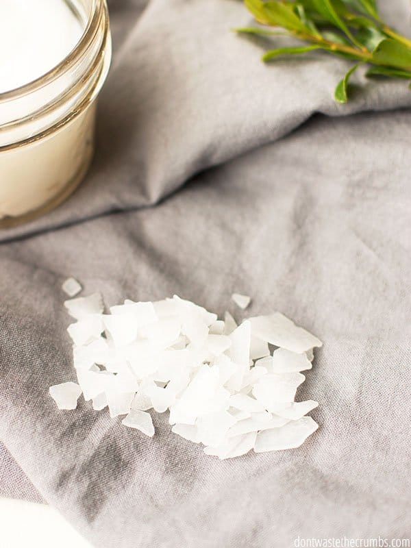 Small, irregularly shaped magnesium flakes lie on a piece of cloth next to a finished jar of the magnesium lotion recipe.