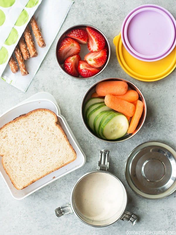 Reusable pouch with pretzel stick, container of strawberries, container of sliced cucumbers and baby carrots, and container with sandwich.