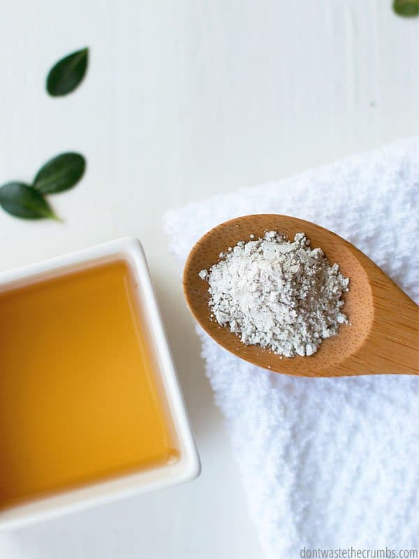 Pictured is a wooden spoon holding powdered bentonite clay on top of a white folded bath towel. A small square dish of apple cider vinegar sits to the left of the spoon.