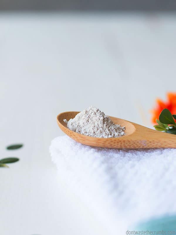 A wooden spoon holding bentonite clay powder sits on top of a fresh white towel that is neatly folded.