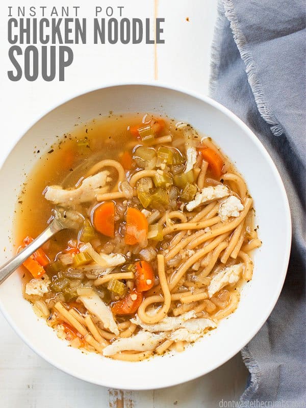 The cover of our Instant Pot Chicken Noodle Soup. A full bowl of soup with a spoon in it sitting on a white table, next to a blue napkin. 