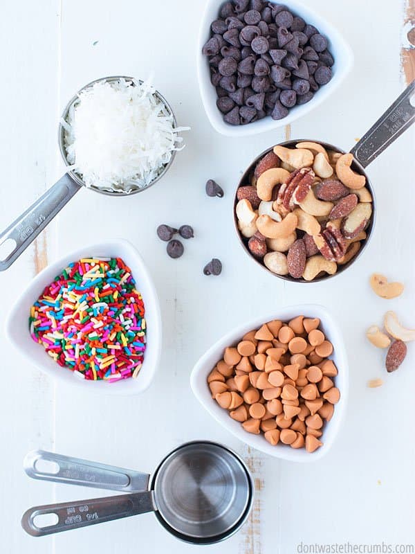 A measuring cups of shredded coconut, a small bowl of chocolate chips, a measuring cups of assorted nuts, a small bowl of peanut butter chips, two empty measuring cups stacked, and a small bowl of rainbow sprinkles on a table.