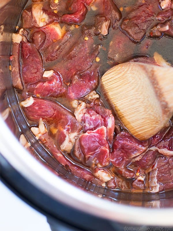 Raw chunks of beef with garlic and seasonings in an Asian sauce inside the Instant Pot, being stirred with a wooden spoon.