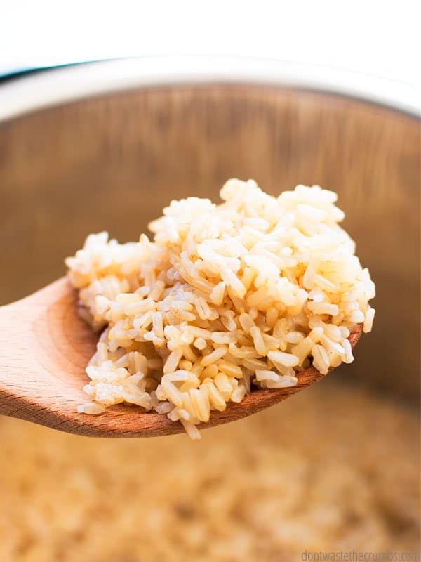 An up close view of a wooden spoon with a spoonful of brown rice on it. In the background is the Instant Pot.