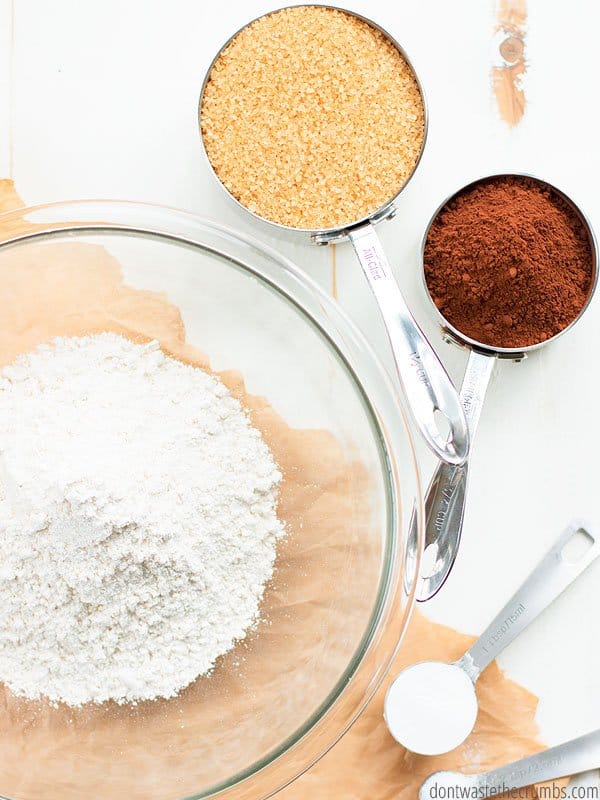 Let's start making this tasty homemade chocolate cake mix recipe! Pictured is flour in a large glass mixing bowl. In separate measuring spoons there is Dutch cocoa, sugar, baking powder, and salt.