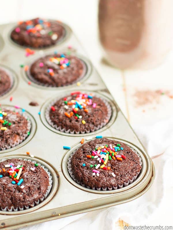 This homemade chocolate cake mix recipe makes the most delicious cupcakes! Pictured is a muffin tin with baked chocolate cupcakes in liners and decorated with a dash of sprinkles.