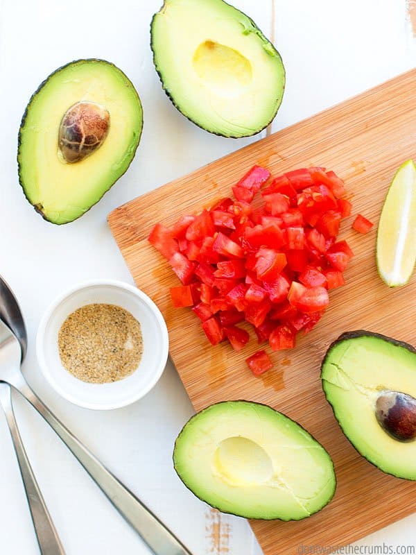Sliced avocados and diced tomatoes on a wooden cutting board with a spoon and fork beside the cutting board.