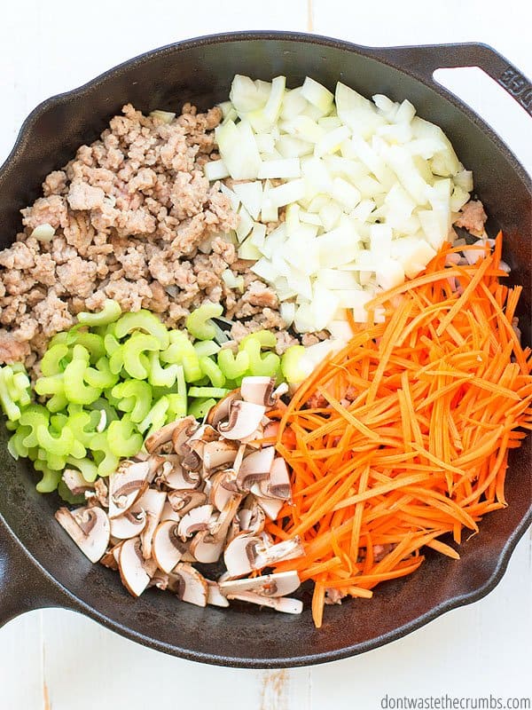 A skillet is filled with ground pork, onions, celery, mushrooms, and shredded carrots.
