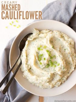 Stylish round white serving bowl filled with creamy mashed cauliflower, garnished with diced green onions. Spoons alongside ready to dig in. Text overlay Creamy Mashed Cauliflower.