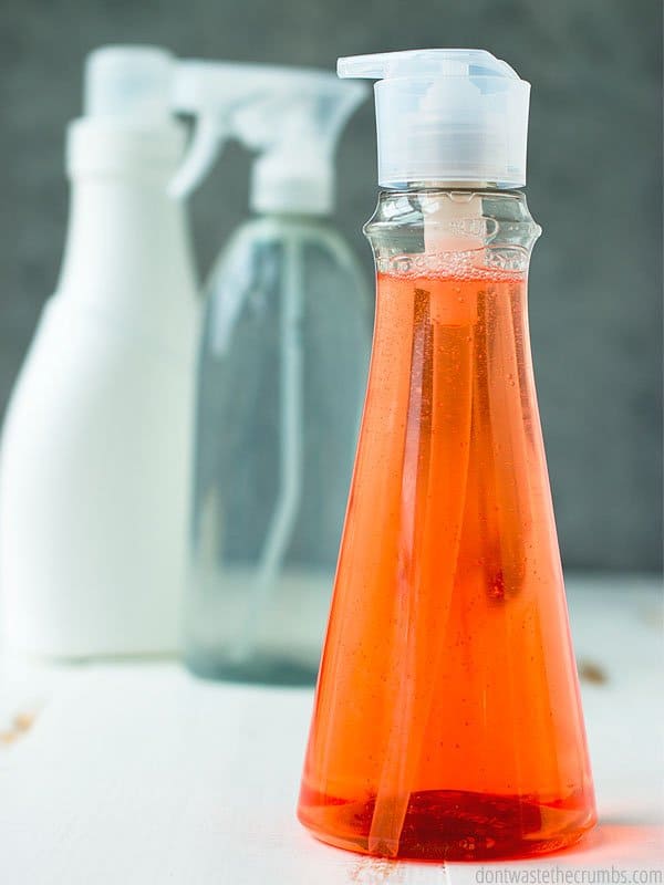 Three plastic dispenser bottles, one clear with red liquid, one clear with green liquid and one a solid white. All on a distressed wood table. Text overlay 8 Harmful & Toxic Household Cleaners to Avoid. 