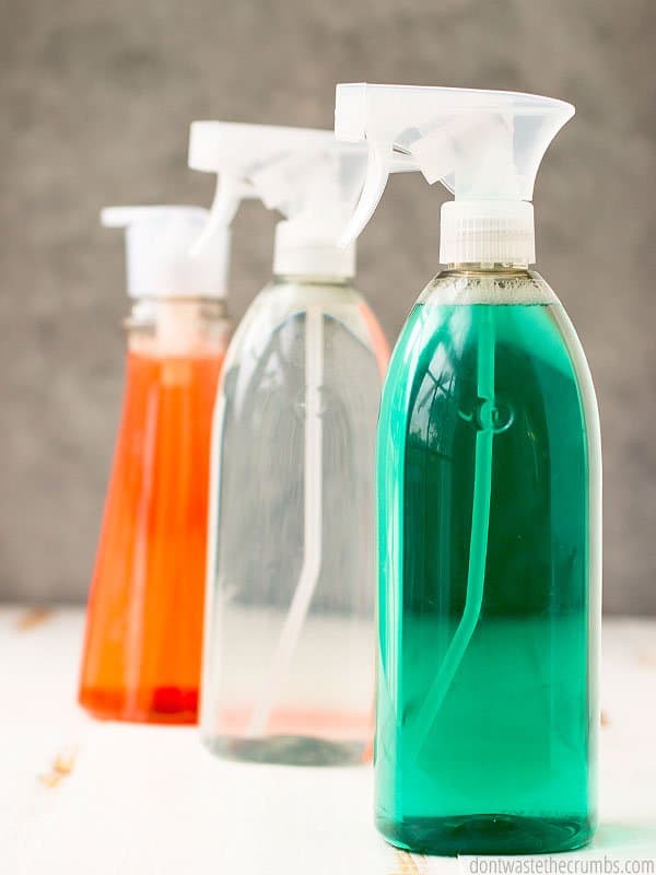 Three plastic dispenser bottles, one clear with red liquid, one clear with green liquid and one a solid white. All on a distressed wood table. Text overlay 8 Harmful & Toxic Household Cleaners to Avoid.