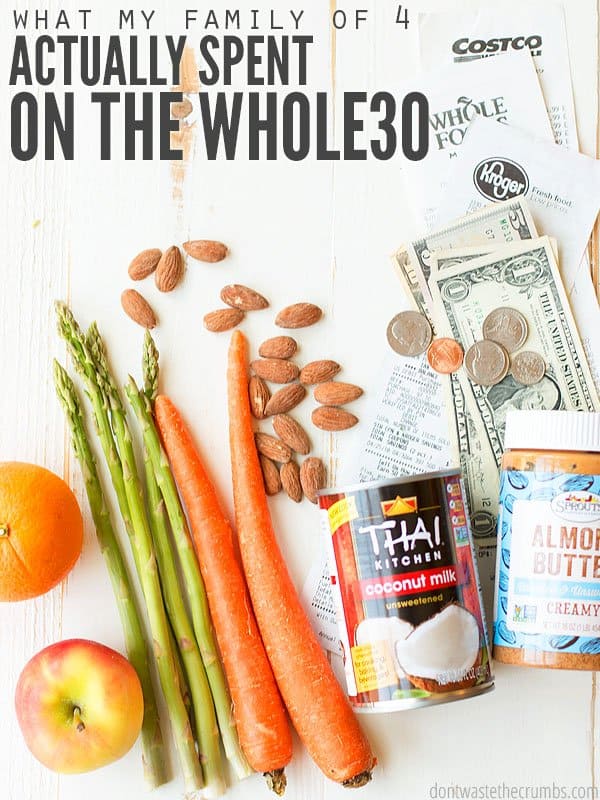 Fresh carrots, asparagus spears, an orange, apple, almonds, a can of coconut milk and a jar of almond butter, with a grocery store receipt and dollar bills, all sitting on a whitewashed wooden kitchen table. Text overlay What My Family of 4 Actually Spent on the Whole30.