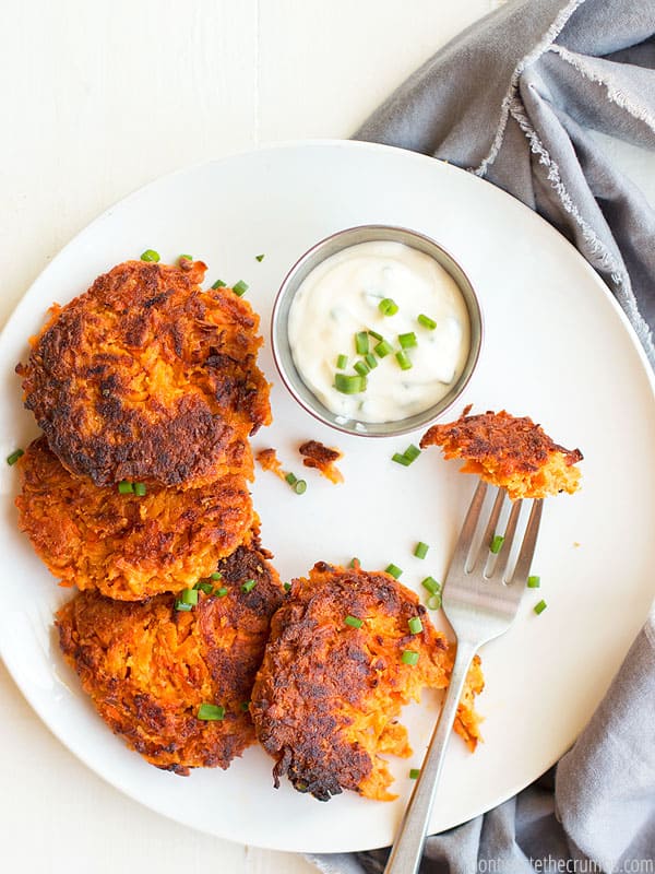 White dinner plate with four round, fried Sweet Potato fritters, one with a bit take out of it. A small dish of dipping sauce sits on the side. Text overlay Carrot Sweet Potato Fritters.