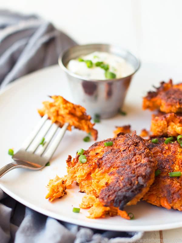 White dinner plate with four round, fried Sweet Potato fritters, one with a bit take out of it. A small dish of dipping sauce sits on the side. Text overlay Carrot Sweet Potato Fritters.