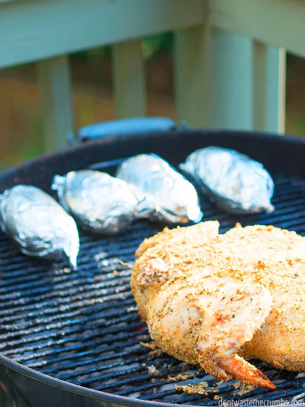 Whole seasoned chicken on round a BBQ grill with a silver set of tongs. Steam rises in the background with two potatoes wrapped in aluminum foil in the foreground. Text overlay One Week Grilling Meal Plan.