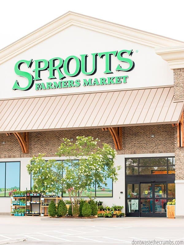 Green Sprouts Farmers Market basket sitting outside with trees in the background. Text overlay 15 Ways to Save Money at Sprouts.
