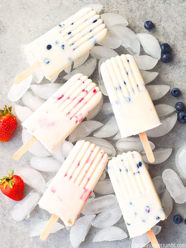 Five blueberry and strawberry yogurt popsicles sit on top of a bed of ice, with a garnish of fresh strawberries and blueberries. 