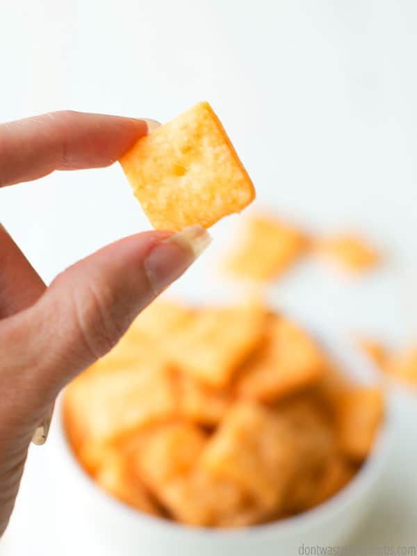 Hand holding a homemade cheez-it with a bowl more cheez-it crackers.