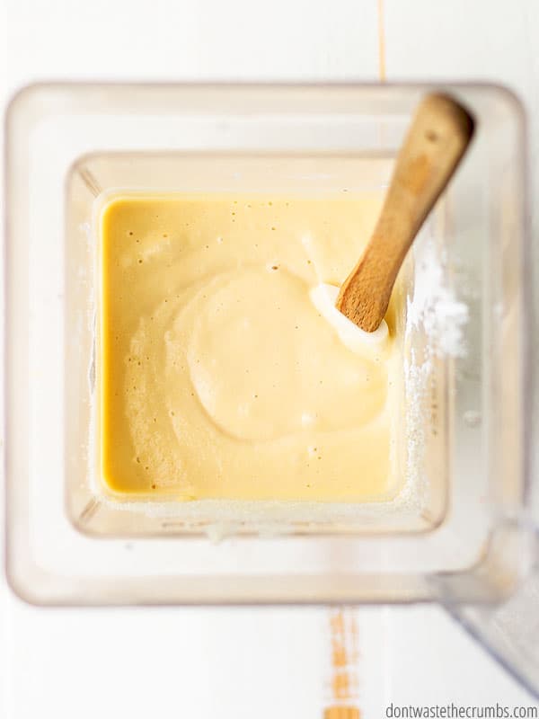 A blender is filled with a rich, brown pancake batter. A wooden spoon is used to make a final stir before pouring the batter.
